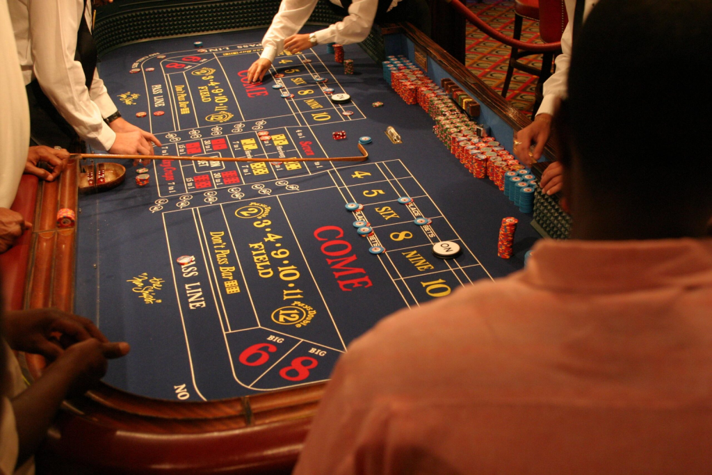 A craps table in a casino, with dealers managing bets and players observing. Chips are stacked on the side, and the table layout includes sections like "Pass Line" and "Come.